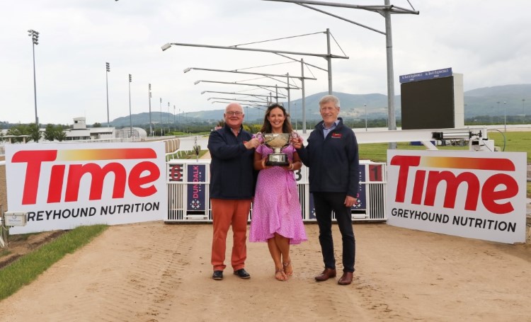 This is an image of three people. A man on the left, a woman in the middle and a man on the right. All three are standing up and holding the trophy for the 2026 Dundalk International. They are standing in front of the traps on the track of Dundalk Stadium. It is a sunny day and the mountains are in the background.