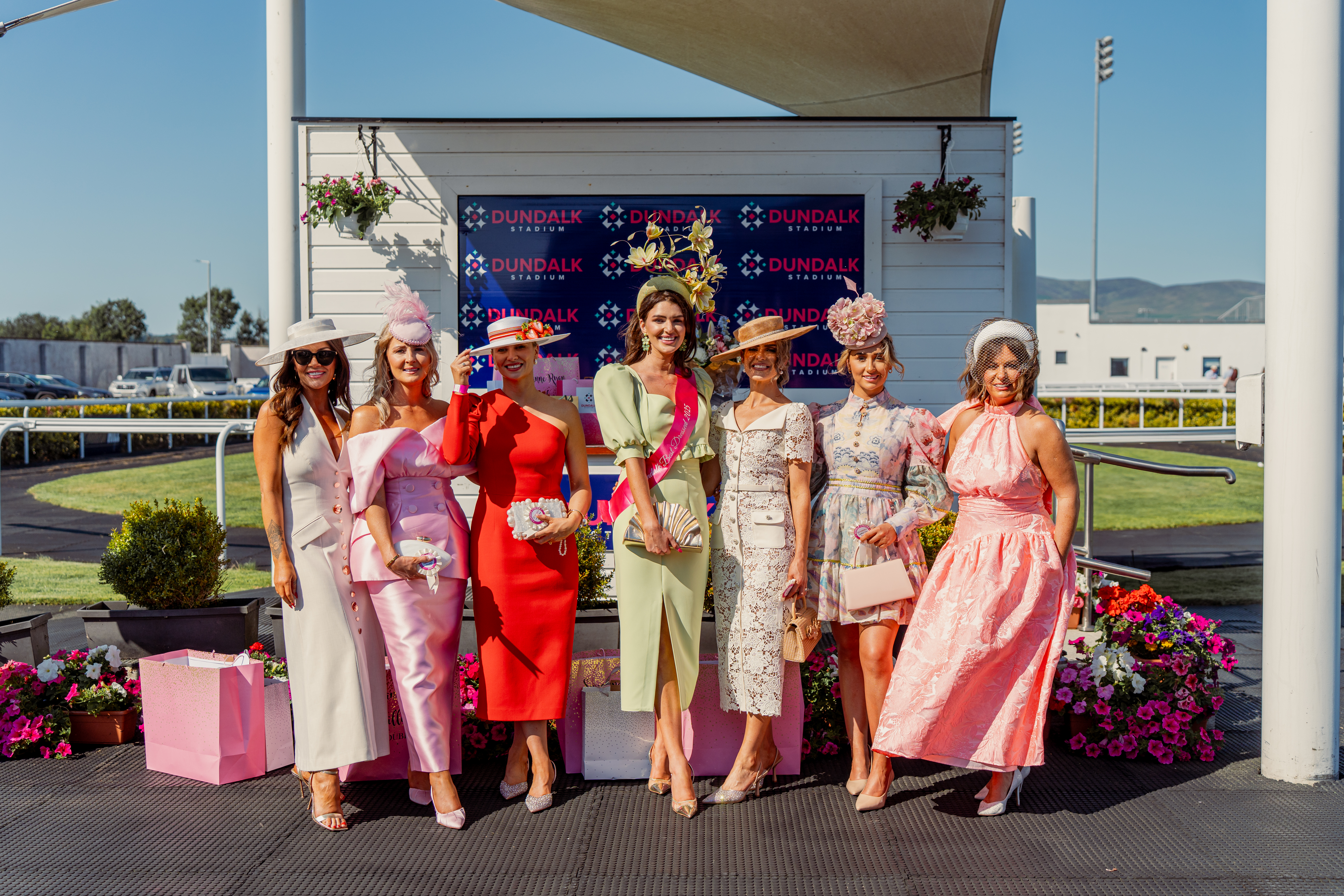 2025 Ladies Day Winner with runners up and judges This is an image of three incredibly dressed women standing outside Dundalk Stadium on a sunny day. These women are the 2026 Ladies Day runners up, judges and winner