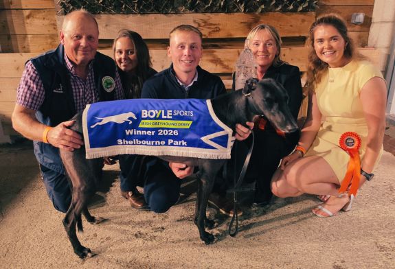 Image shows a family, the Holland family, in front a timber backdrop and kneeling behind a black greyhound, their 2026 BOYLE Sports Irish Greyhound Derby champion, Bockos Gold