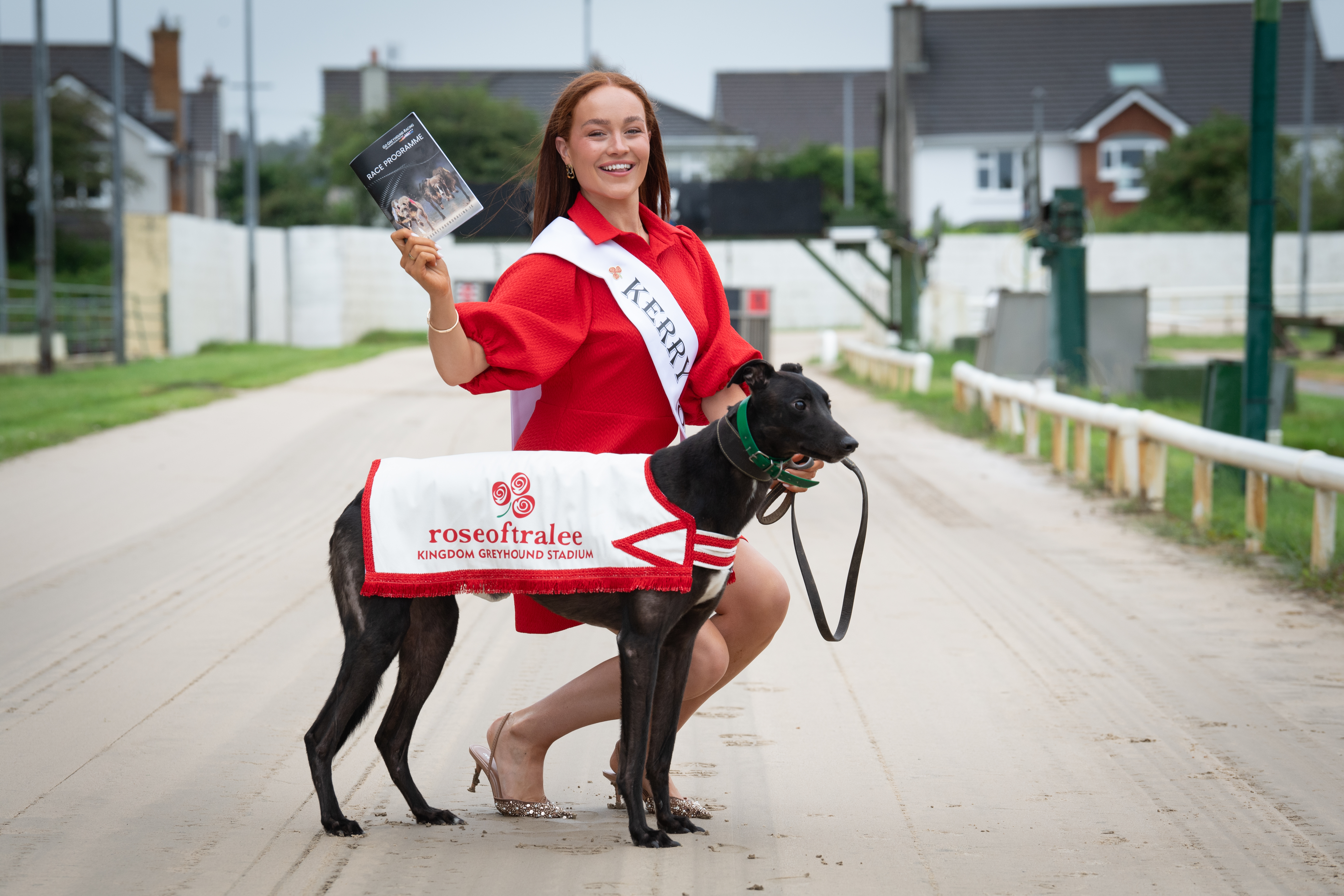 Kerry Rose Laura Daly, pictured at the Kingdom Greyhound Stadium in Tralee with a greyhound to launch the 2025 Afternoon Racing with the Roses event on Saturday 16th August A woman in a red dress, the 2025 Kerry Rose Laura Daly, is pictured on the sand track at the Kingdom Greyhound Stadium in Tralee with a black greyhound wearing a Rose of Tralee branded jacket