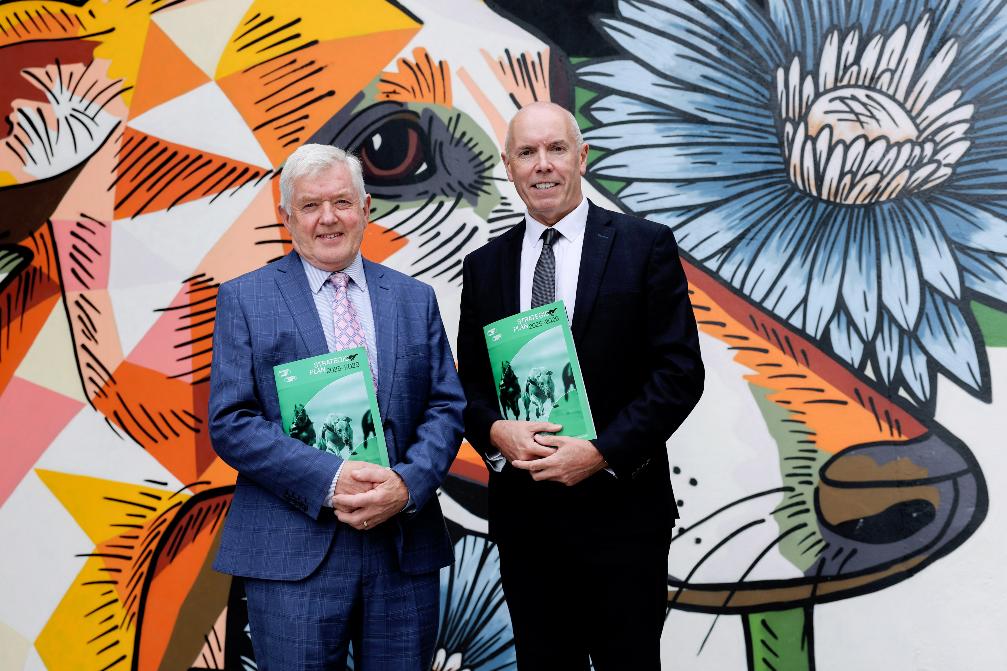 RCÉ/GRI Chairman Pat Flanagan and CEO Tim Lucey launching the Rásaíocht Con Éireann Strategic Plan 2025 - 2029 at Shelbourne Park Greyhound Stadium Picture shows two men, RCÉ/GRI Chairman Pat Flanagan and CEO Tim Lucey, standing in front of colourful mural at Shelbourne Park Greyhound Stadium to launch the new Strategic Plan 2025 - 2029 which they are both holding in their hands.