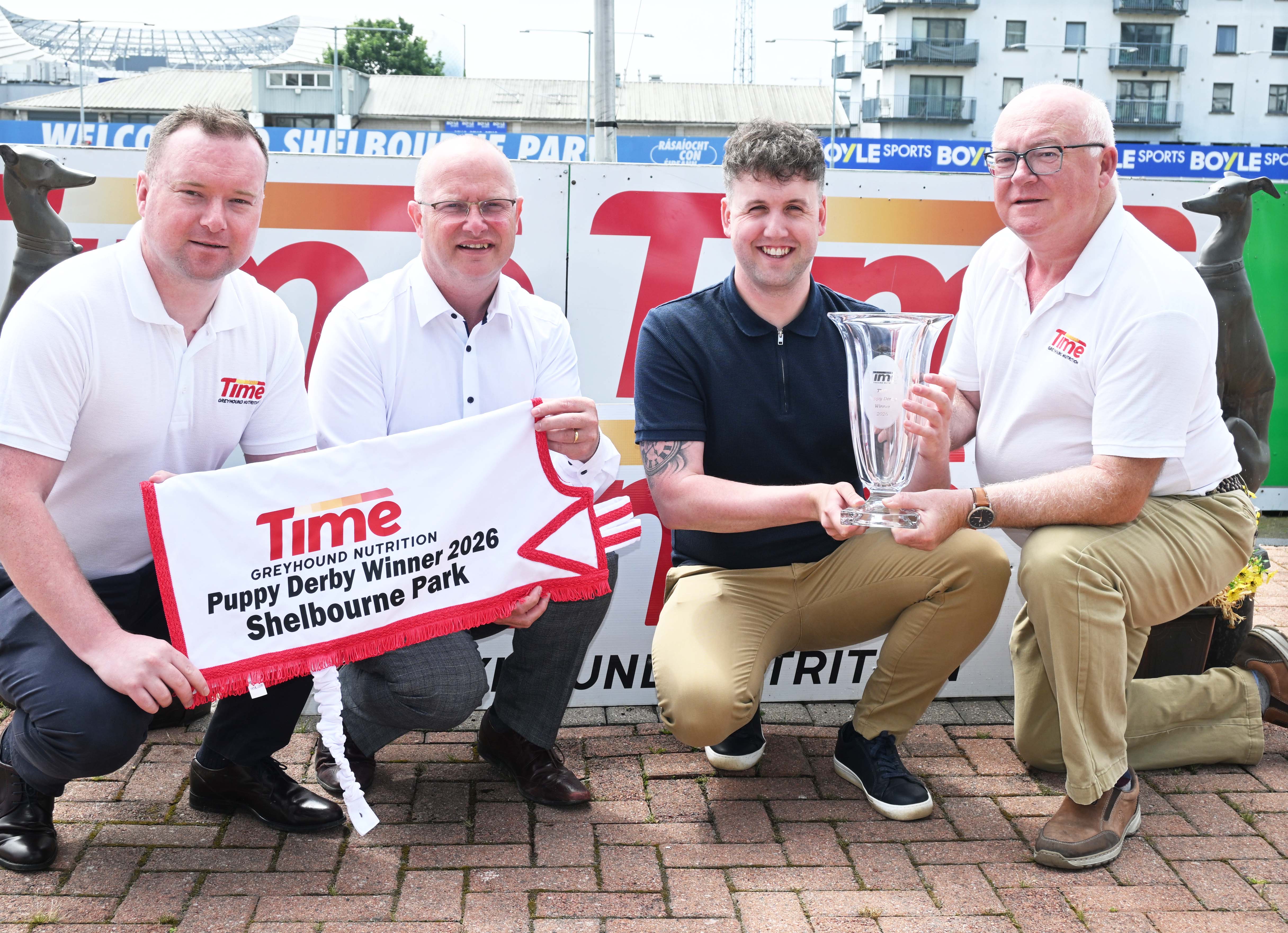 Image shows four men pictured at Shelbourne Park Greyhound Stadium on a sunny day, holding a greyhound presentation sheet to laucn the Time Puppy Derby